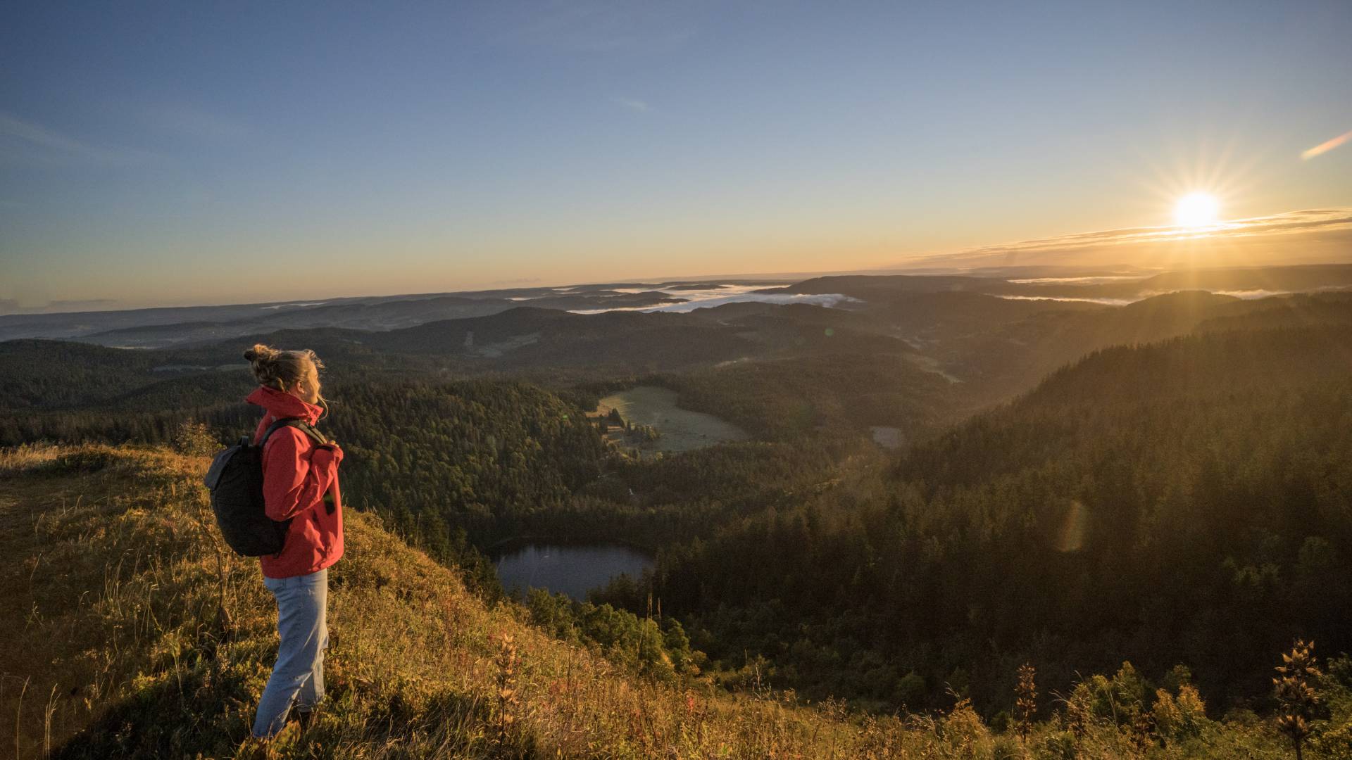 Wanderin steht auf dem Feldberg und schaut auf den Feldsee bei Sonnenaufgang